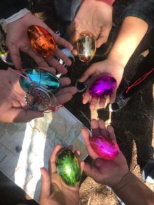 Six people's hands, each of which holding a differently colored metallic egg.